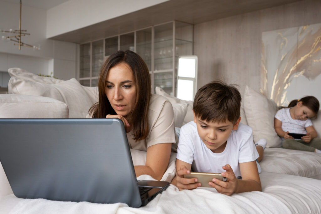 A woman and a child are lying on a bed and using a laptop and a phone